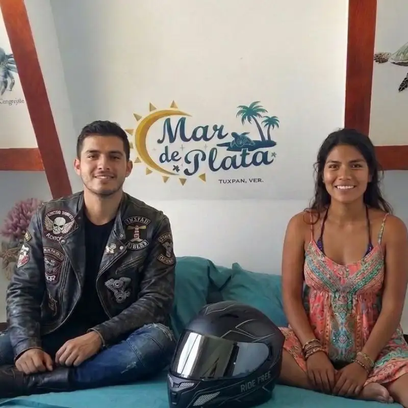 Pareja joven sonriente con casco de moto sentada en una cama frente al logotipo mural de Mar de Plata, Tuxpan, dentro de la Cabaña 2.