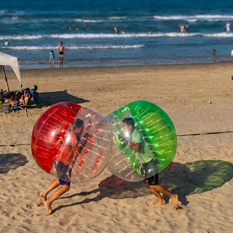 Dos personas jugando y chocando dentro de Bumper Balls (pelotas choconas) gigantes en la arena de la playa Mar de Plata en Tuxpan, con el mar de fondo.