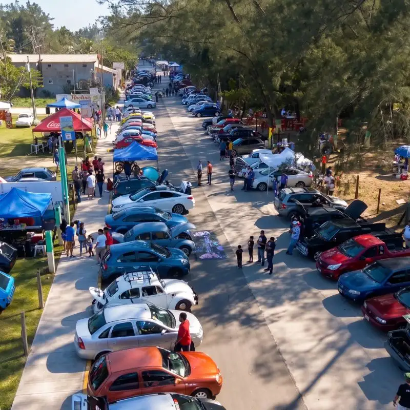 Vista aérea de una gran exhibición de autos tuning y car audio en Tuxpan, con decenas de vehículos modificados estacionados a lo largo de un camino rodeado de árboles y personas admirándolos.