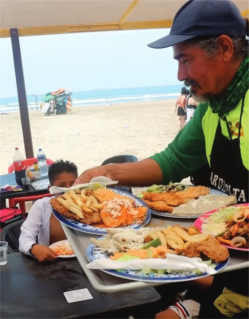 Mesero sirviendo una charola con abundantes platillos tradicionales y mariscos a una familia en las palapas de nuestro restaurante en Tuxpan frente al mar.