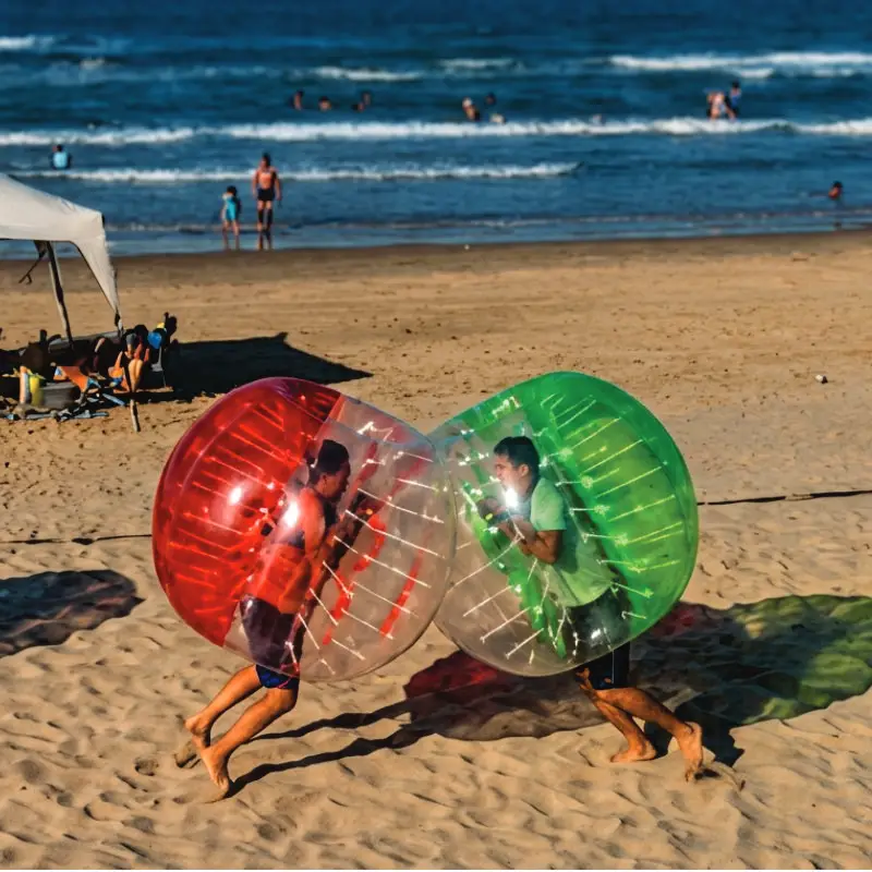 Dos personas riendo y chocando mientras usan grandes Bumper Balls inflables, una roja y una verde, en la arena de la playa de Mar de Plata Tuxpan. Al fondo se ve el Golfo de México, otras personas y una carpa de playa.